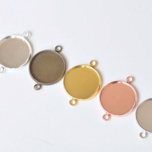 Może przedstawiać: Pięć okrągłych metalowych tacek na zawieszki z trzema pętlami do przymocowania łańcuszka lub sznurka. Tace są srebrne, brązowe, złote, różowe złoto i przezroczyste.