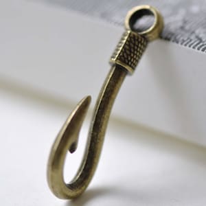 Op de afbeelding: Een gouden metalen haakbedel met een getextureerd ontwerp op het bovenste gedeelte. De haak is gebogen en heeft een scherpe punt.