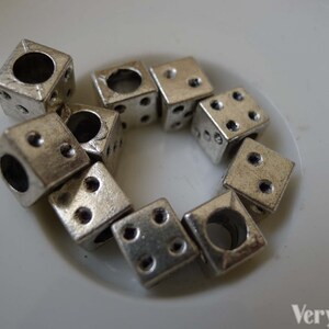 May include: A group of silver metal dice charms with holes in the center. The dice are arranged in a circle on a white surface.