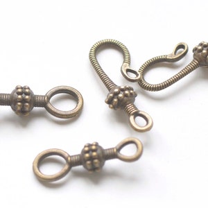 May include: Antique brass-colored jewelry clasps. The clasps have a decorative beaded center and loop ends. One clasp has a hook closure. The clasps are on a white background.
