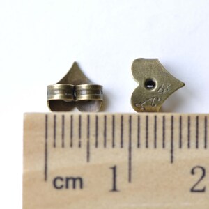 Op de afbeelding: Twee bronzen hartvormige oorbelruggen, ongeveer 1 cm lang, worden naast een liniaal getoond.