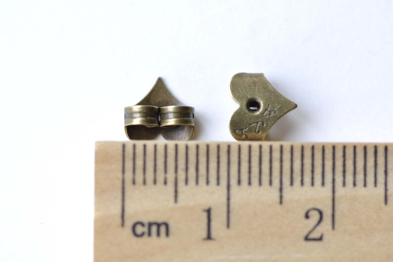 Op de afbeelding: Twee bronzen hartvormige oorbelruggen, ongeveer 1 cm lang, worden naast een liniaal getoond.