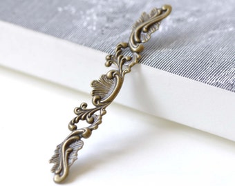 May include: A decorative metal embellishment with a scroll design in an antique brass finish. The embellishment is shaped like a long, thin rectangle with intricate details.