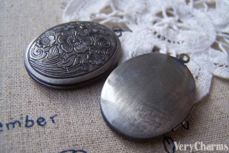 May include: Two silver oval locket pendants. One pendant has a floral design and the other is plain. Both pendants have a loop for attaching a chain.