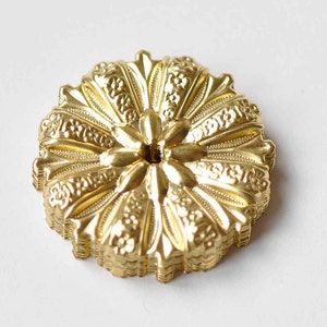 May include: A gold-colored metal decorative embellishment with a floral design. The embellishment has a circular shape and a raised center with a small hole.