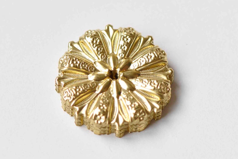 May include: A gold-colored metal decorative embellishment with a floral design. The embellishment has a circular shape and a raised center with a small hole.