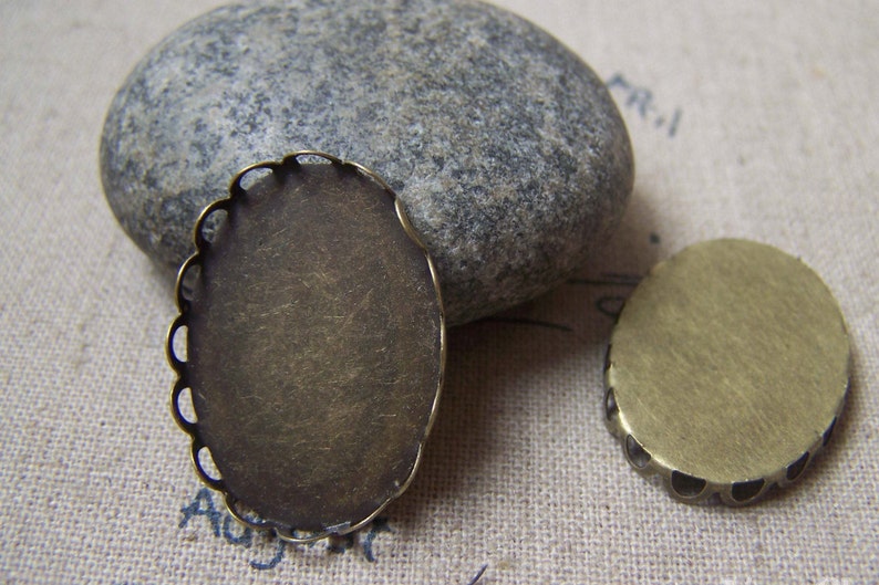 May include: Two oval-shaped metal settings for jewellery making. One setting has a scalloped edge and the other has a smooth edge. Both settings are a brass colour.