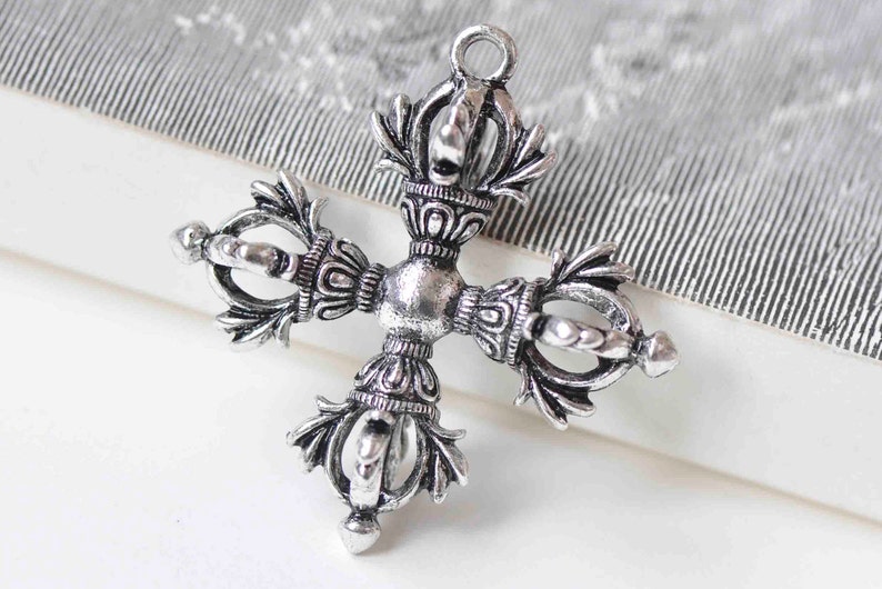 May include: Silver-toned metal pendant in the shape of a cross with intricate details. The cross is adorned with four ornate, stylized figures, each with a circular center and a pointed top.