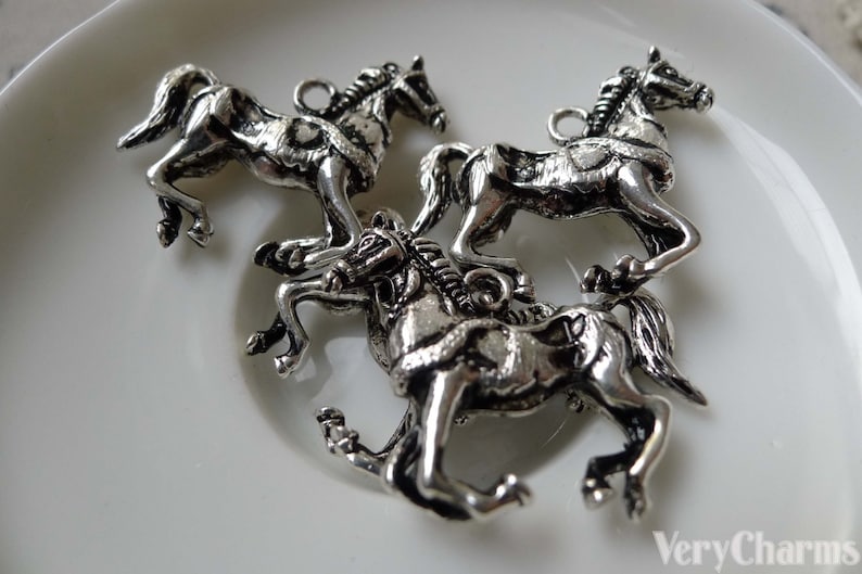 May include: Three silver-colored horse charms in various poses, each with a loop for attaching to jewelry or crafts. The horses are detailed with manes, tails, and harnesses, and are displayed on a white surface.