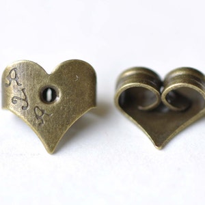 May include: A pair of heart-shaped earring backs in a brass finish. The backs are designed to hold earrings in place. The backs are engraved with the word "AYA".