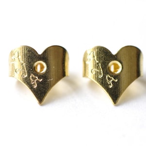 Puede incluir: Un par de respaldos de pendientes de corazón de oro con un pequeño agujero en el centro. Los respaldos están grabados con un diseño.
