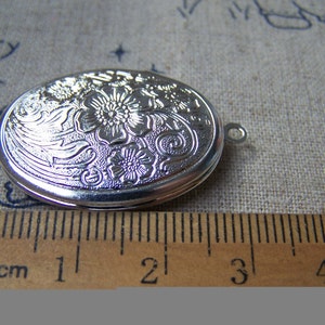 K&ouml;nnte beinhalten: Silbernes ovales Medaillon mit floralem Design. Das Medaillon ist etwa 2 cm lang.