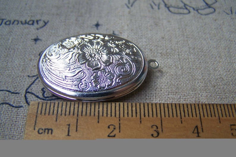 K&ouml;nnte beinhalten: Silbernes ovales Medaillon mit floralem Design. Das Medaillon ist etwa 2 cm lang.
