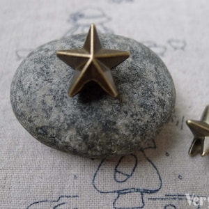 Könnte beinhalten: Zwei antike goldfarbene Metall-Stern-Anhänger. Der größere Stern liegt auf einem grauen Stein, der kleinere Stern auf einem weißen Stoffhintergrund.