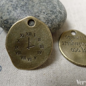 Puede incluir: Dos abalorios de metal de color dorado antiguo. Un abalorio es redondo y presenta números romanos alrededor del borde y un diseño de espada en el centro. El otro abalorio es redondo y presenta el texto "no. 1 is the greatest gift" en una fuente cursiva.