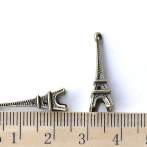 May include: Two antique gold Eiffel Tower charms, one with a loop at the top and the other with a loop at the bottom, measured against a ruler in inches.