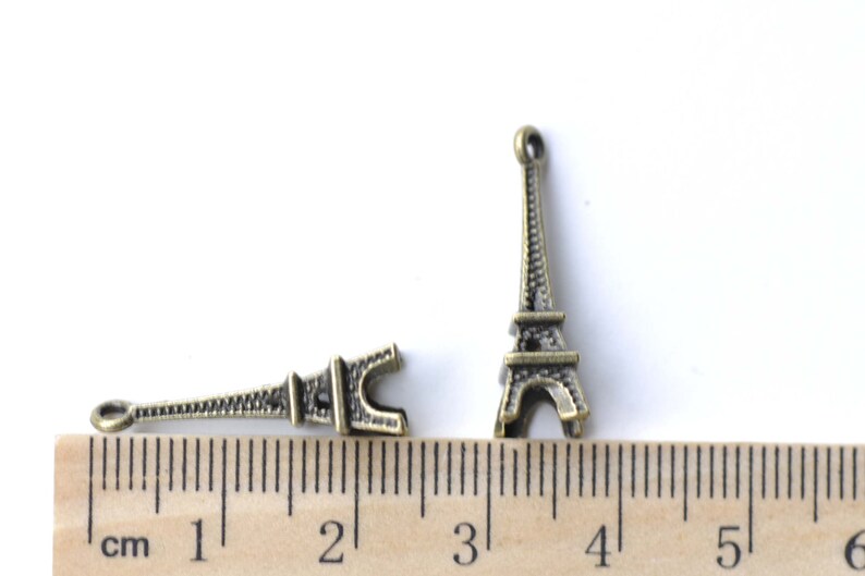 May include: Two antique gold Eiffel Tower charms, one with a loop at the top and the other with a loop at the bottom, measured against a ruler in inches.