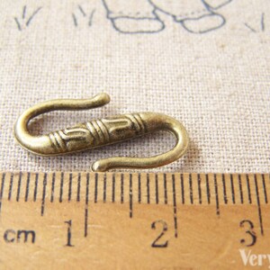 May include: A gold-toned metal hook clasp with an ornate design. The hook is approximately 2 cm long and is shown next to a ruler.