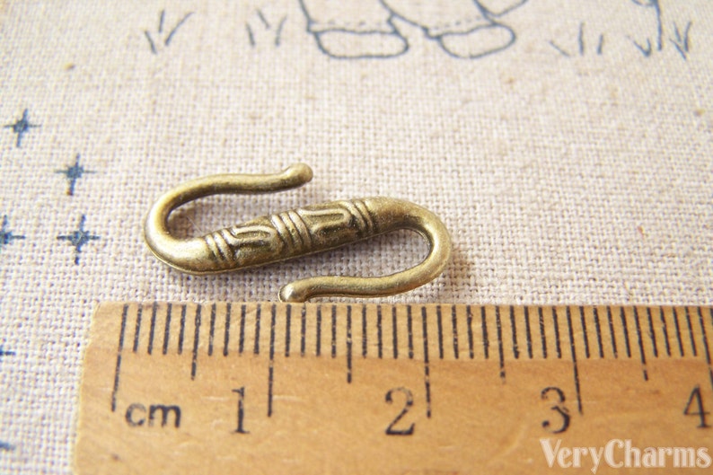 May include: A gold-toned metal hook clasp with an ornate design. The hook is approximately 2 cm long and is shown next to a ruler.