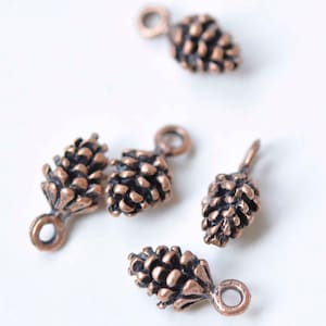 Könnte beinhalten: Nahaufnahme von mehreren kleinen, bronzefarbenen Tannenzapfen-Anhängern. Jeder Anhänger hat eine detaillierte, strukturierte Oberfläche und eine kleine Schlaufe zum Befestigen an Schmuck oder anderen Bastelarbeiten. Die Anhänger sind auf einer weißen Oberfläche verstreut.