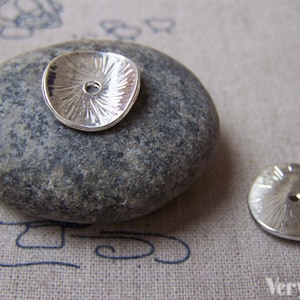 May include: Two silver metal round charms with a textured surface and a hole in the centre. The charms are on a grey stone and a beige fabric background.