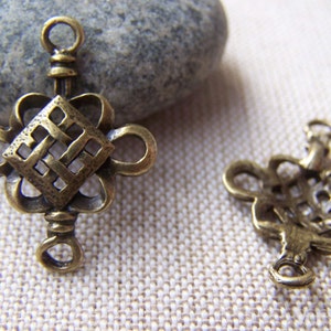 May include: Two antique gold-toned metal charms in the shape of a Chinese knot, also known as a lucky knot. Each charm has a loop at the top for attaching to a necklace, bracelet, or other jewelry.