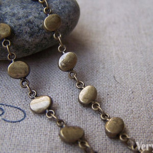 May include: A close-up of a gold chain with round, flat, metal beads. The chain is lying on a textured fabric surface.