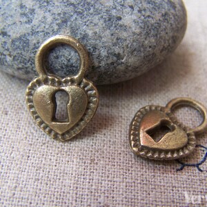 May include: Two antique gold heart-shaped lock charms with keyholes. The charms are on a light brown fabric background.