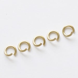 Könnte beinhalten: Sechs goldfarbene Jump Rings, auch bekannt als Splitt-Ringe, sind in einer Reihe auf einer weißen Oberfläche angeordnet. Diese Ringe werden häufig in der Schmuckherstellung und im Handwerk verwendet.