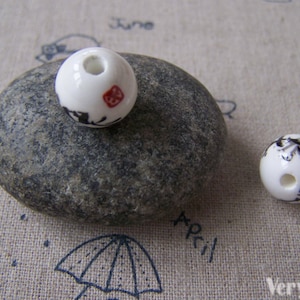 May include: Two white ceramic beads with black and red painted designs. The beads have a hole in the center for stringing.