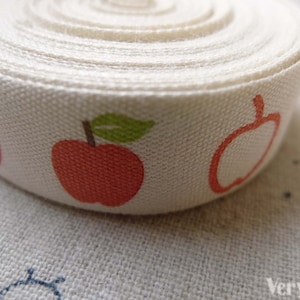 May include: White cotton ribbon with a repeating pattern of red apples and green leaves.