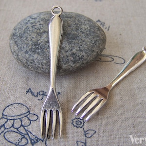 Op de afbeelding: Twee zilverkleurige vorkcharms met een lus aan de bovenkant om aan sieraden of andere knutselwerken te bevestigen. De vorken liggen op een textuurrijke stoffen achtergrond.