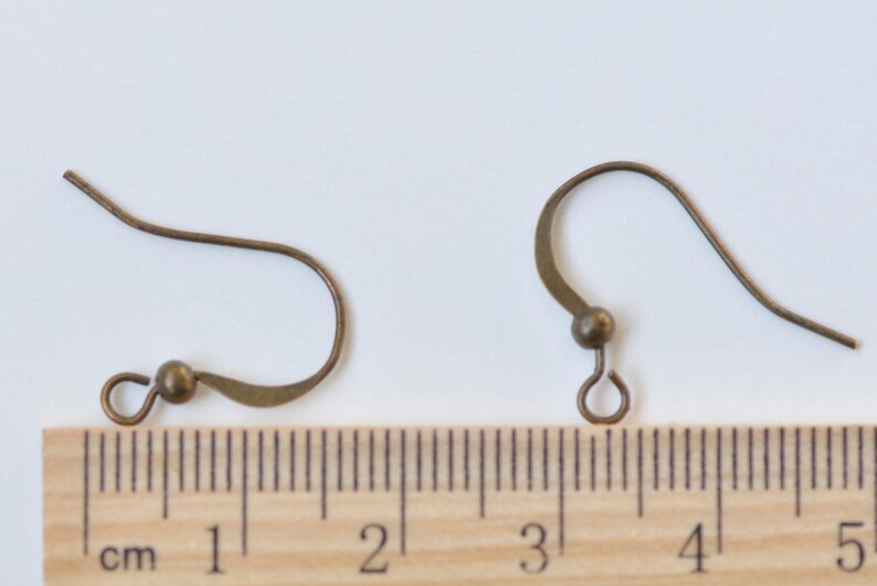 May include: Two bronze-colored earring hooks with a small loop at the end. The hooks are about 2 cm long and are shown next to a ruler.