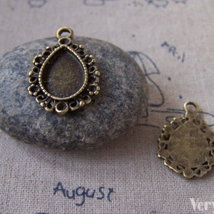May include: Two antique gold-toned metal pendant charms with ornate designs. One charm features a teardrop-shaped frame with a clear glass insert. The other charm has a round shape with a detailed pattern.