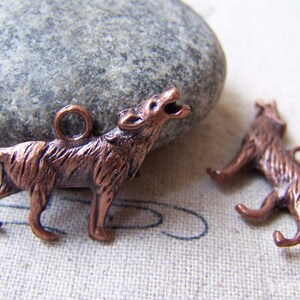 May include: Two copper-colored wolf charms. One wolf is howling with its mouth open, and the other is in a running pose. Both charms have a small loop at the top for attaching to jewelry or other crafts. The background includes a gray stone and a patterned fabric.