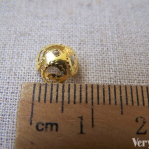 May include: Gold metal bead with a decorative openwork design. The bead is approximately 1 cm in diameter and has a hole for threading.