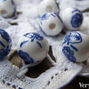 Puede incluir: Cuentas de cer&aacute;mica blanca con dise&ntilde;os florales azules. Las cuentas son redondas y tienen un agujero en el centro. Est&aacute;n dispuestas sobre una superficie de crochet blanco.