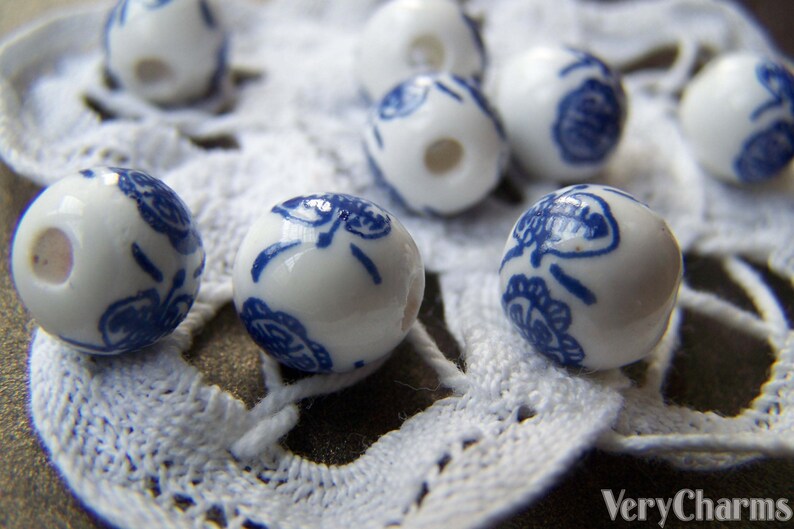 Puede incluir: Cuentas de cer&aacute;mica blanca con dise&ntilde;os florales azules. Las cuentas son redondas y tienen un agujero en el centro. Est&aacute;n dispuestas sobre una superficie de crochet blanco.
