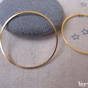 Può includere: Due cerchi in metallo dorati, uno più grande dell'altro, giacciono su una superficie di tessuto beige. Il cerchio più grande è appoggiato su una pietra grigia.