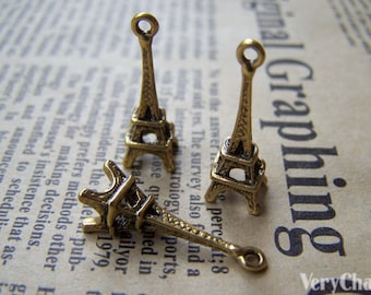 May include: Three gold-toned Eiffel Tower charms, each with a loop at the top for attaching to a necklace or bracelet. The charms are on a brown paper background with the text "VeryCharms" and "Graphing" printed on it.
