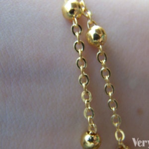 May include: Close-up of a gold-colored bracelet with two strands. Each strand features small, round beads and delicate chain links. The jewelry is displayed against a blurred background.
