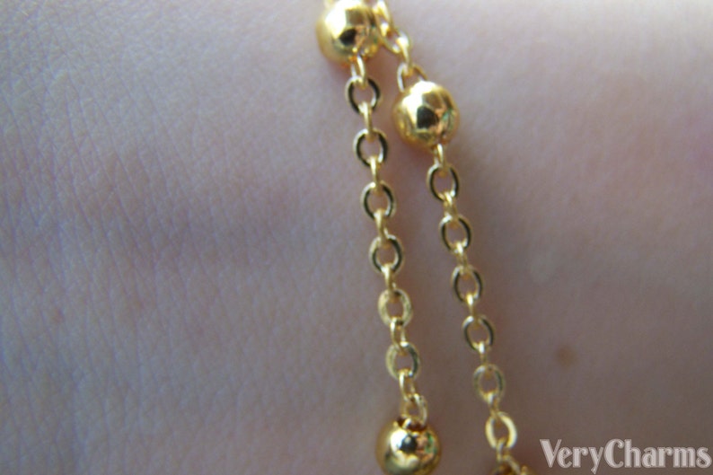 May include: Close-up of a gold-colored bracelet with two strands. Each strand features small, round beads and delicate chain links. The jewelry is displayed against a blurred background.