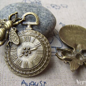May include: Two antique bronze charms, one featuring a bee perched on a clock face and the other a bee with wings outstretched. Both charms have a detailed, intricate design.