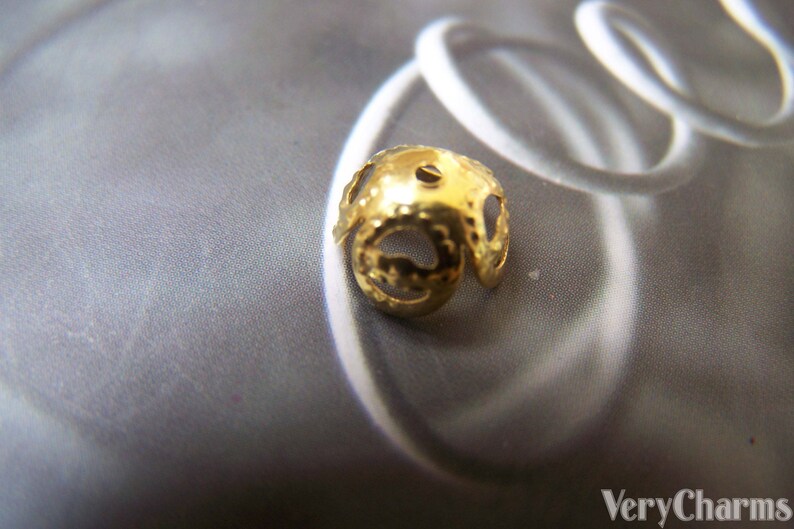 May include: Gold-toned metal bead with a decorative, openwork design. The bead has a round shape and a small hole in the center.