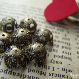 May include: A group of eight antique gold-toned metal beads with a decorative design. The beads have a raised pattern of small dots and a scalloped edge. The beads are arranged on a textured surface.