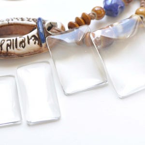 May include: Clear, rectangular glass pendants with rounded corners. The pendants are displayed with a beaded necklace featuring brown, blue, and tan beads. A ceramic pendant with script is also visible.