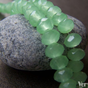May include: A strand of light green faceted glass beads, arranged in a double row, resting on a gray stone. The beads are smooth and translucent, with a slight shimmer.