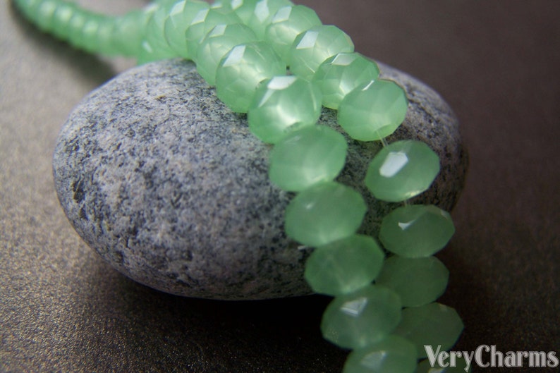 May include: A strand of light green faceted glass beads, arranged in a double row, resting on a gray stone. The beads are smooth and translucent, with a slight shimmer.