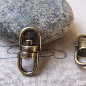 May include: Two antique gold swivel clasps, also known as trigger snaps, are shown on a textured fabric surface. The clasps are oval-shaped with a small, round, swiveling mechanism in the center.
