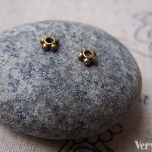Può includere: Due piccole perline di metallo dorate con un design a forma di fiore. Le perline sono su una roccia grigia.